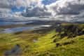 Lush green hills and lakes under a cloudy sky, with distant mountains and sunlight breaking through clouds.