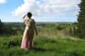 Woman in floral dress standing in grassy field, looking at a scenic landscape with trees and clouds.