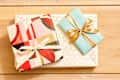 Overhead shot of three wrapped gifts - a large rectangular gift wrapped in white and gold patterned paper, a small book sized package wrapped in a light blue paper with gold ribbon , and on the left side is a square gift wrapped in gold, pink and red abstract paper.