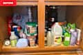 Cluttered kitchen cabinet under the sink filled with cleaning supplies, bottles, and plastic bags.