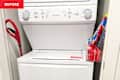 Washer and dryer stacked in a laundry nook with a red shovel leaning against the wall and laundry supplies in a clear container.