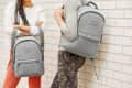Two people with gray backpacks leaning against a brick wall, one wearing camo pants.