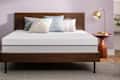 Modern bedroom with a wooden bed frame, white mattress, assorted pillows, and a gold lamp on a wooden side table.