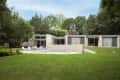 Modern house with large glass windows, surrounded by trees, featuring a pool and lounge chairs on a grassy lawn.