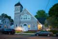 Historic church building with arched windows and a tower, illuminated at dusk, with parked cars in front.