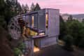 Modern metal-clad house with large windows, surrounded by trees, featuring a unique outdoor sculpture at sunset.
