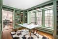 Home office with green built-in bookshelves, large windows, white desk, and checkered rug, leading to a colorful living room.