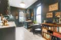 Cozy kitchen with dark green cabinets, round table, bookshelves, and framed art on walls.