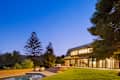 Modern house exterior at dusk with large windows, pool, and landscaped yard featuring trees and lounge chairs.