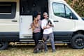 A woman holding an orange cat and a man with a black cat stand by a camper van, with a brown dog sitting in front.