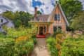 Charming orange cottage with steep roof, surrounded by lush garden and vibrant flowers, under a partly cloudy sky.