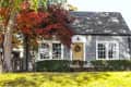 Gray shingle cottage with yellow door, autumn foliage, and manicured shrubs.