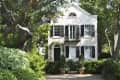 Two-story house with black shutters, arched windows, and lush greenery in the front yard.