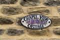 Oval sign reading "Chapel View Cottage" with purple flowers on a stone wall.