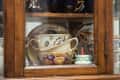 Colorful teacups and saucers displayed in a wooden cabinet, featuring floral designs and a small decorative candle.