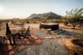 Desert landscape with a hot tub, wooden chairs, and a chiminea on a stone patio, with mountains in the background.