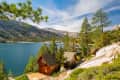 Lakeside view with wooden cabins, pine trees, and mountains under a blue sky with wispy clouds.
