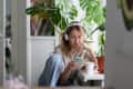 A woman with headphones sits on the floor, looking at her phone, with a cup and croissant on a table surrounded by plants.