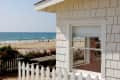 Beachfront cottage with white picket fence, red flowers, and ocean view. People walking on sandy beach in the background.