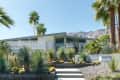 Mid-century modern house with geometric facade, surrounded by cacti and palm trees, set against a mountain backdrop.