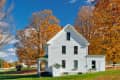 White farmhouse surrounded by vibrant autumn trees with orange leaves under a clear blue sky.
