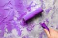 Hand using a paint roller to apply purple paint on a textured gray wall.