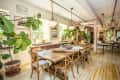 Rustic dining room with wooden table, chairs, large potted plants, and chandeliers, surrounded by framed art and windows.