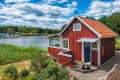 Red wooden house by a calm lake, surrounded by greenery and boats in the distance under a blue sky with clouds.