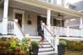 Woman in business attire walking down steps of a large porch with white railings and potted plants.