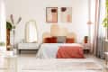 Modern bedroom with a scalloped headboard, abstract art, and plants. White dresser, round mirror, and coral bedding.