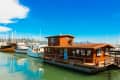 Houseboat with wooden exterior docked in a marina, surrounded by sailboats under a clear blue sky.