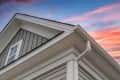 Roofline of a house featuring gray siding, a white window, and a downspout against a colorful sunset sky.