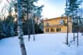 Modern wooden house in snowy forest, surrounded by tall trees.