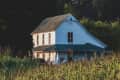 Two-story white farmhouse with a green metal roof, surrounded by tall grass and plants.