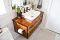Wooden bathroom vanity with white sink, black faucet, potted plant, and toiletries on a tiled wall background.