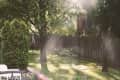 Sunlit backyard with green grass, trees, a swing set, and a wooden fence, featuring blooming flowers in the foreground.