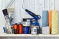 Paint supplies on a shelf, including brushes, rollers, paint cans, and a caulking gun against a wooden wall.