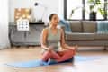 Woman meditating on a blue yoga mat in a living room, wearing headphones, with a sofa and desk in the background.