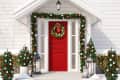 Red front door with Christmas wreath, garlands, and small decorated trees, flanked by lanterns on a snowy porch.