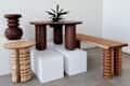 Wooden furniture pieces including a round table with a plant, a stool, and a bench, displayed on white pedestals.
