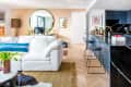 Modern living room with white sofa, blue pillow, large mirror, and open kitchen with black marble countertop and bar stools.