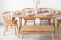 Dining table set with wooden chairs and bench, pink table runner, white dishes, and a vase with branches.