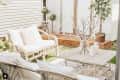 Cozy patio with wicker furniture, white cushions, wooden table, potted plants, and string lights near a white shed.