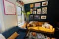 Cozy living room with a yellow sofa, blue couch, gallery wall, and a tall plant by a window with a lamp and books.