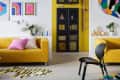 yellow black and white squiggly shaped rug with line striped tiger like pattern, yellow couch, pink and pink and white striped pillow, black wood chair, black glass book case, colorful graphic art, white floating shelf behind couch, plants, colorful splat patterned rug
