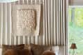 curtain wall with hanging fiber art,brown round throw pillows, beige linens, wood panel ceiling,