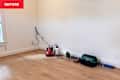 Empty room corner with light wood flooring, tools including a red sander, green laser level, and a black tool bag.