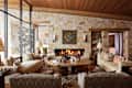 Cozy living room with a stone fireplace, circular coffee table, patterned rugs, and large windows showing snowy outdoors.