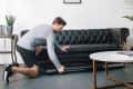 Man in gray shirt and shorts kneeling, pulling a black treadmill from under a tufted leather sofa in a modern living room.