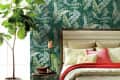 Bedroom with tropical leaf wallpaper, beige bedding, red throw, and a potted plant beside a round side table.